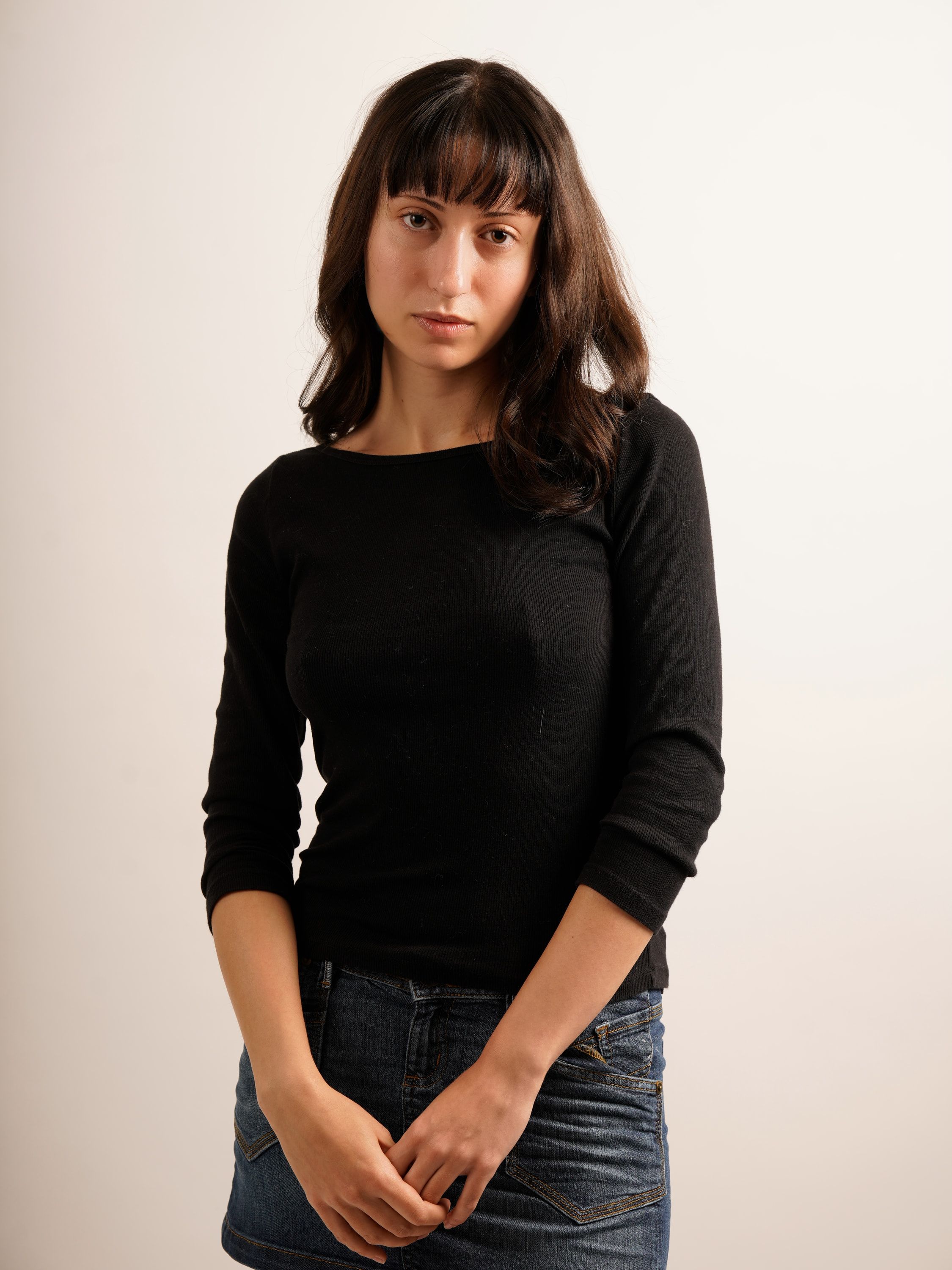 Colour digital of Chrissa facing the camera, hands clasped, in a black top and denim mini skirt.