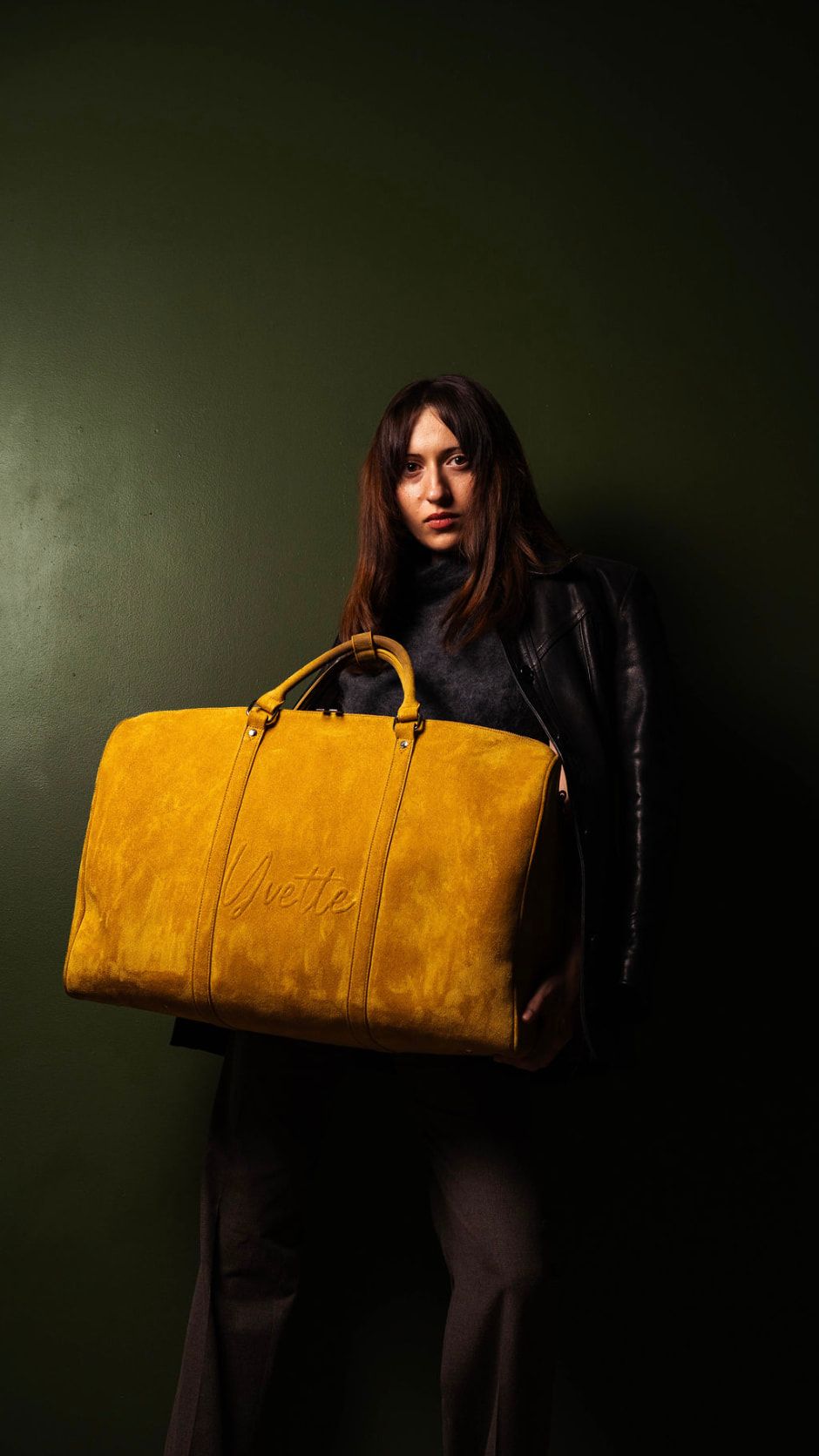 Chrissa holding a large ochre suede duffel bag against her hip, looking into the camera, in a black leather jacket by a dark green wall.